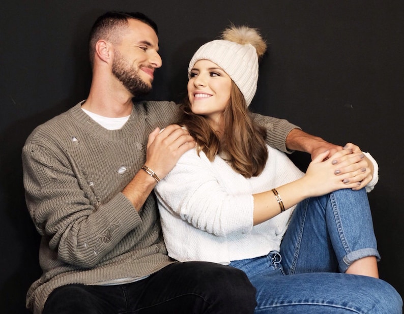 May include: A couple sitting together, the woman is wearing a white knit hat with a fluffy pom pom and a white sweater, the man is wearing a gray sweater with holes in it.