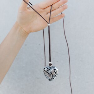 May include: A silver heart-shaped pendant with an intricate filigree design hangs from a brown leather cord.