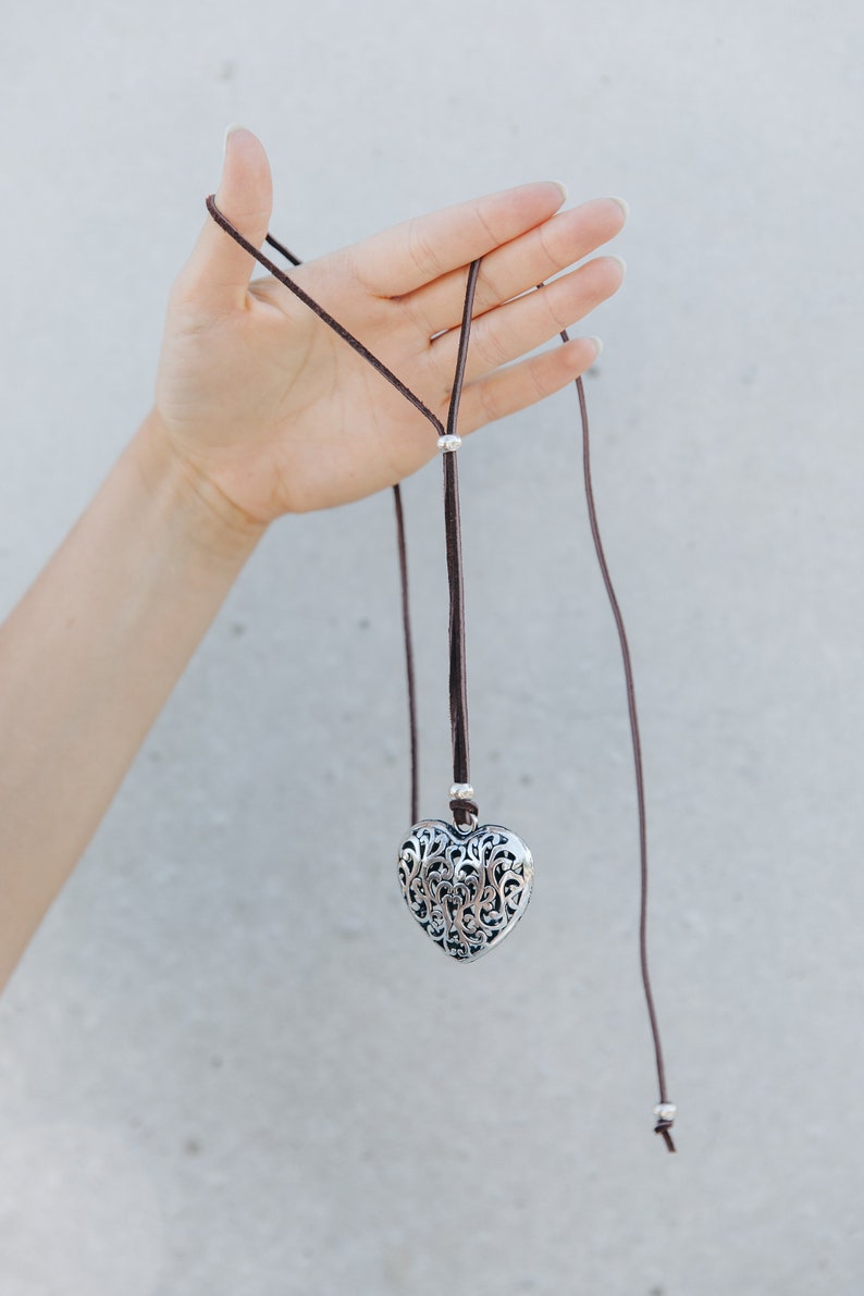 May include: A silver heart-shaped pendant with an intricate filigree design hangs from a brown leather cord.