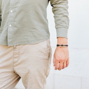 May include: A man wearing a light green button-down shirt and beige pants. He is wearing a brown leather bracelet with a silver buckle on his right wrist.