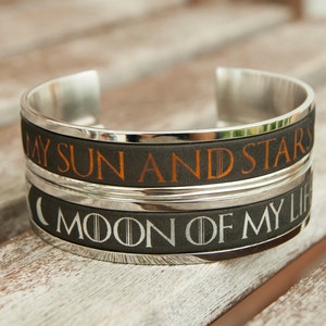 Könnte beinhalten: Ein silbernes Armreif-Armband mit zwei Bändern. Das obere Band trägt den Text "My Sun and Stars" in orangefarbener Schrift auf schwarzem Hintergrund. Das untere Band trägt den Text "Moon of My Life" in weißer Schrift auf schwarzem Hintergrund.