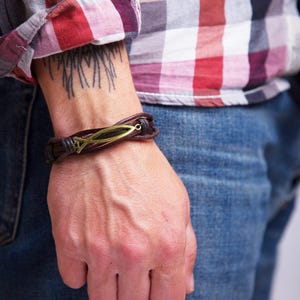 Puede incluir: Un brazalete de cuero marrón con un dije de pescado de metal. El brazalete se lleva en la muñeca.
