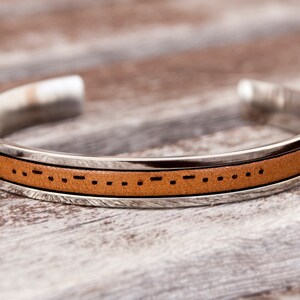 A leather cuff bracelet with a stainless steel band with Morse code engraved. The bracelet is shown on a light brown wooden surface. Stainless steel adjustable bracelet with custom morse code engraved makes meaningful long distance relationship gift for boyfriend or girlfriend.