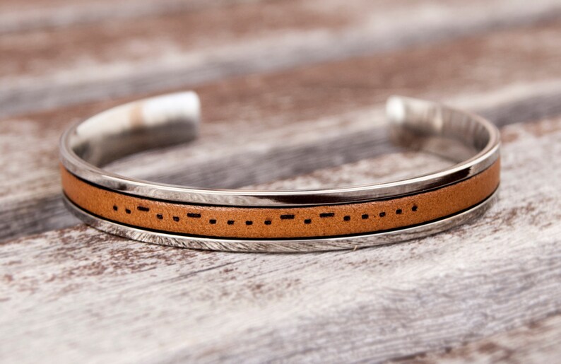 A leather cuff bracelet with a stainless steel band with Morse code engraved. The bracelet is shown on a light brown wooden surface. Stainless steel adjustable bracelet with custom morse code engraved makes meaningful long distance relationship gift for boyfriend or girlfriend.
