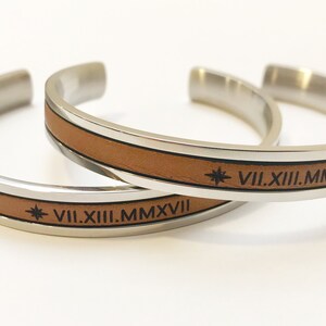 May include: Two silver bracelets with brown leather bands. Each bracelet has a star and the Roman numeral date VII.XIII.MMXVII engraved on the leather.