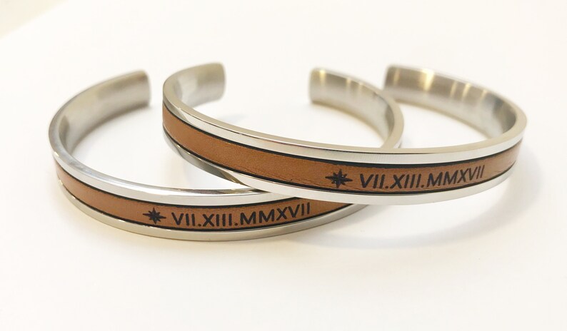 May include: Two silver bracelets with brown leather bands. Each bracelet has a star and the Roman numeral date VII.XIII.MMXVII engraved on the leather.