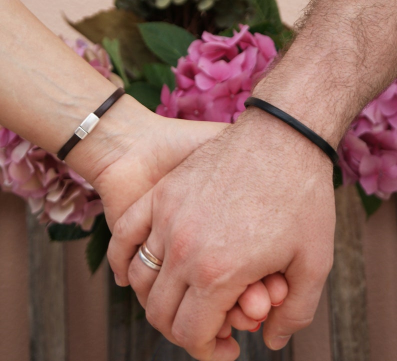 May include: A close-up of two hands holding each other. The person on the left is wearing a brown leather bracelet with a silver clasp. The person on the right is wearing a black leather bracelet. Both hands are wearing rings.