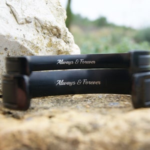 Op de afbeelding: Twee zwarte metalen armbanden, elk gegraveerd met de inscriptie "Always & Forever".