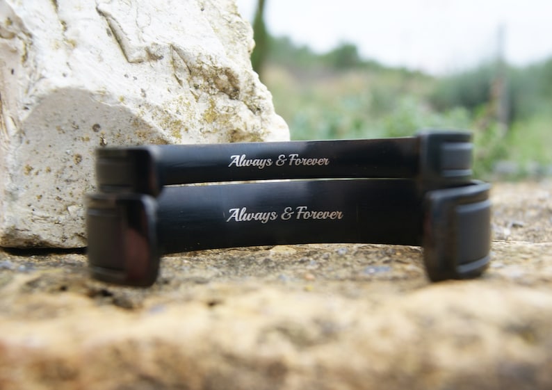Op de afbeelding: Twee zwarte metalen armbanden, elk gegraveerd met de inscriptie "Always & Forever".