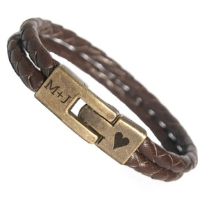 Puede incluir: Pulsera de cuero marrón trenzado con cierre color bronce. El cierre está grabado con las iniciales "M+J" y un símbolo de corazón. La pulsera presenta dos hebras paralelas de cuero trenzado.