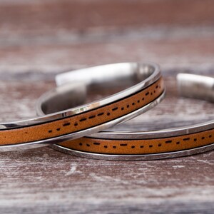 Close-up of two brown leather and stainless steel cuff bracelets. Each bracelet features custom engraved morse code message. A leather cuff bracelet with a stainless steel band with Morse code engraved. The bracelet is shown on a light brown wooden surface. Stainless steel adjustable bracelet with custom morse code engraved makes meaningful long distance relationship gift for boyfriend or girlfriend.