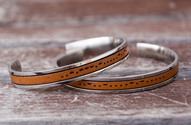 Close-up of two brown leather and stainless steel cuff bracelets. Each bracelet features custom engraved morse code message. A leather cuff bracelet with a stainless steel band with Morse code engraved. The bracelet is shown on a light brown wooden surface. Stainless steel adjustable bracelet with custom morse code engraved makes meaningful long distance relationship gift for boyfriend or girlfriend.