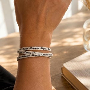 Puede incluir: Una pulsera envolvente blanca con texto negro que repite las palabras Loved, Redeemed, Forgiven, Adopted, Known y Chosen. La pulsera est&aacute; dise&ntilde;ada para envolverse varias veces alrededor de la mu&ntilde;eca, mostrando las palabras en una fuente clara.
