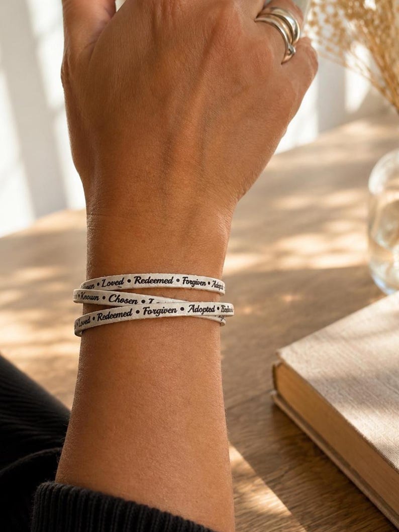 Puede incluir: Una pulsera envolvente blanca con texto negro que repite las palabras Loved, Redeemed, Forgiven, Adopted, Known y Chosen. La pulsera est&aacute; dise&ntilde;ada para envolverse varias veces alrededor de la mu&ntilde;eca, mostrando las palabras en una fuente clara.