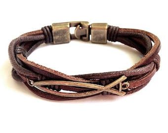 Puede incluir: Un brazalete de cuero marrón con un diseño trenzado y un dije de pez de metal.