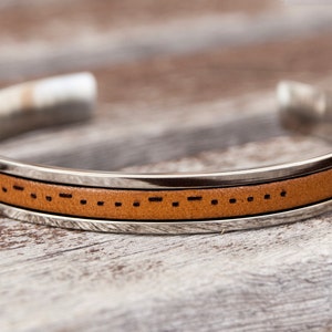 Könnte beinhalten: Ein braunes Lederarmband mit einem silbernen Band und einem Muster aus Punkten und Strichen. Das Armband liegt auf einer Holzoberfläche.
