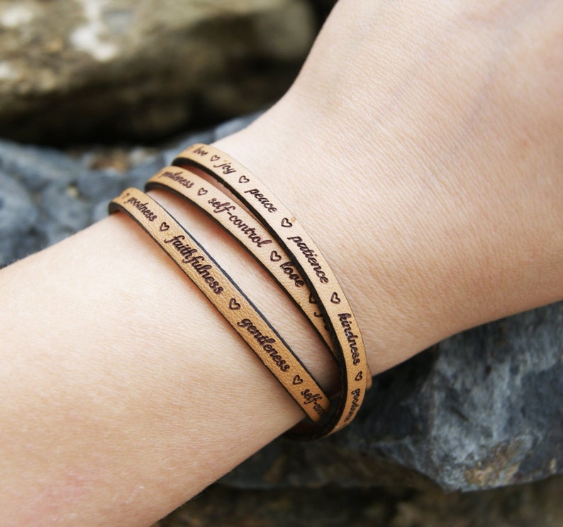 Puede incluir: Un brazalete de cuero de triple vuelta con las palabras "amor, alegr&iacute;a, paz, paciencia, bondad, bondad, fidelidad, gentileza, autocontrol" grabadas en &eacute;l. Cada palabra est&aacute; separada por un s&iacute;mbolo de coraz&oacute;n.