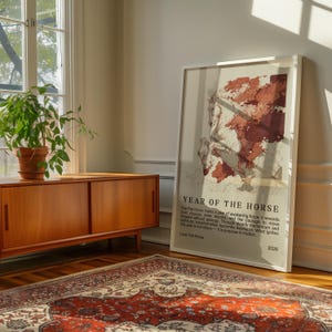 May include: A framed poster with the text "YEAR OF THE HORSE" and a map-like illustration in shades of red and beige. The poster leans against a wall in a room with a wooden cabinet, a potted plant, and a patterned rug.