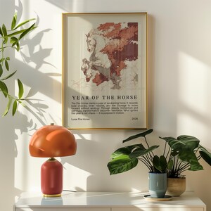 May include: A framed art print with a gold frame featuring a horse illustration with the text "YEAR OF THE HORSE". A red and orange table lamp sits on a white surface. Green plants in pots are also visible.