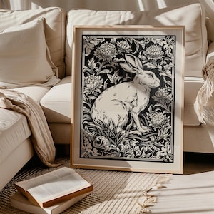 Puede incluir: Una impresión artística en blanco y negro enmarcada que presenta una ilustración detallada de un conejo rodeado de patrones florales. La obra de arte se muestra en un marco de madera de color claro, agregando un toque de elegancia al diseño. La impresión se establece sobre un fondo neutro.