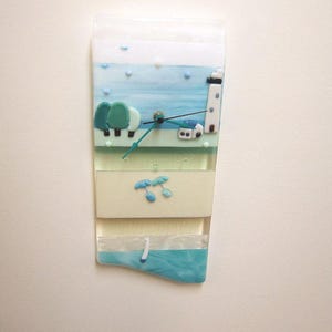 May include: A rectangular wall clock with a coastal scene. The clock face features a lighthouse, trees, and a sailboat, all in shades of blue, green, and white. The clock hands are black, and the overall design is artistic and decorative.