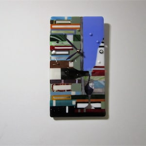 May include: A rectangular fused glass wall clock with a wine theme. The clock features a wine bottle pouring into a glass, a lighthouse, and a blue sky. The clock is made of various colored glass pieces.