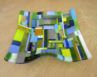 May include: A decorative glass dish with a mosaic design. The dish features a variety of colors, including blue, green, yellow, and brown, arranged in a geometric pattern. The dish has a wavy shape and is displayed on a wooden surface.
