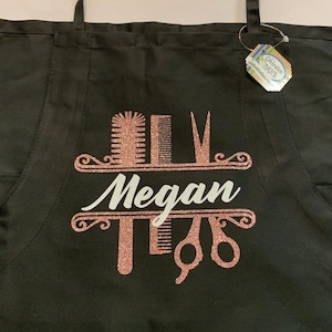 K&ouml;nnte beinhalten: Schwarze Sch&uuml;rze mit einem ros&eacute;goldenen Glitzerdesign mit Kamm, Schere und Haarb&uuml;rste. Der Name "Megan" ist in wei&szlig;er Schrift dargestellt. Die Sch&uuml;rze hat schwarze Tr&auml;ger.