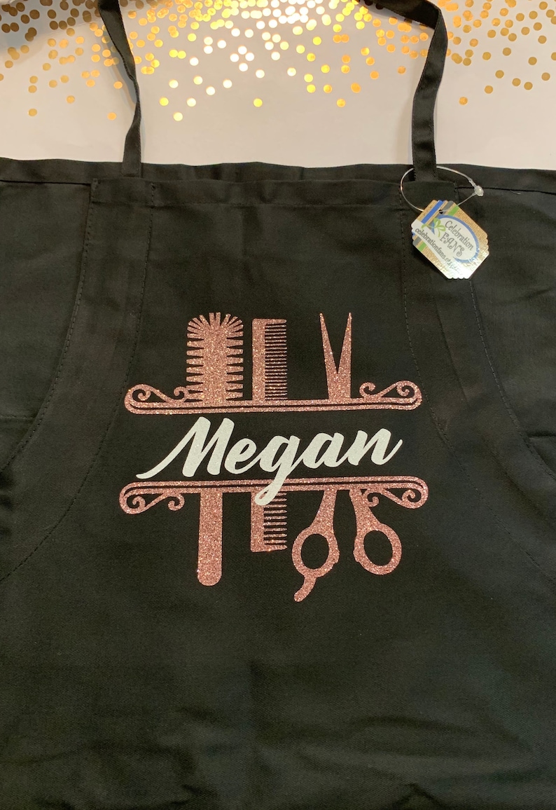 K&ouml;nnte beinhalten: Schwarze Sch&uuml;rze mit einem ros&eacute;goldenen Glitzerdesign mit Kamm, Schere und Haarb&uuml;rste. Der Name "Megan" ist in wei&szlig;er Schrift dargestellt. Die Sch&uuml;rze hat schwarze Tr&auml;ger.