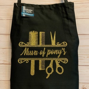 K&ouml;nnte beinhalten: Schwarze Sch&uuml;rze mit goldenem Glitzerdesign mit K&auml;mmen, Scheren und dem Text "Mura of pony's". Die Sch&uuml;rze hat ein Etikett mit der Aufschrift "Celebration Fans".