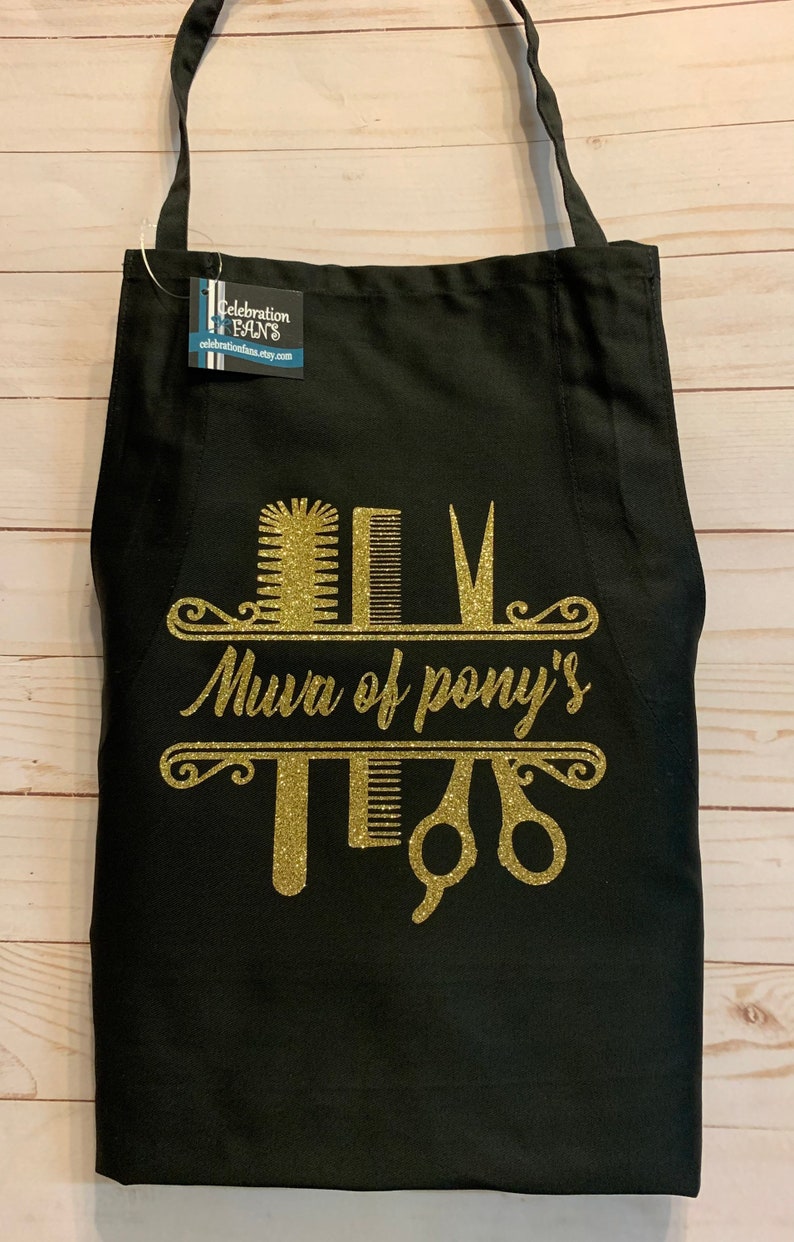K&ouml;nnte beinhalten: Schwarze Sch&uuml;rze mit goldenem Glitzerdesign mit K&auml;mmen, Scheren und dem Text "Mura of pony's". Die Sch&uuml;rze hat ein Etikett mit der Aufschrift "Celebration Fans".