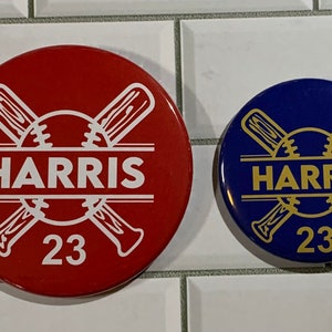 May include: Two round baseball themed buttons with the name "HARRIS" and the number "23" on them. The button on the left is red with white lettering and the button on the right is blue with gold lettering.
