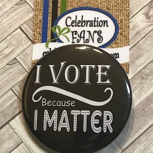 May include: A black button with white text that reads "I VOTE Because I MATTER." The button is round and glossy. Behind it is a card with the words "Celebration FANS" and a website address.