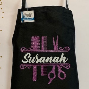 K&ouml;nnte beinhalten: Schwarze Sch&uuml;rze mit einem glitzernden lila Design mit K&auml;mmen, Scheren und dem Namen "Susanah" in wei&szlig;er Schrift. Die Sch&uuml;rze hat einen Nackenriemen und ein Etikett.