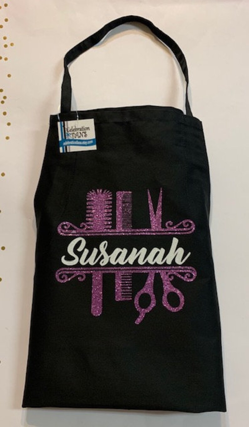K&ouml;nnte beinhalten: Schwarze Sch&uuml;rze mit einem glitzernden lila Design mit K&auml;mmen, Scheren und dem Namen "Susanah" in wei&szlig;er Schrift. Die Sch&uuml;rze hat einen Nackenriemen und ein Etikett.