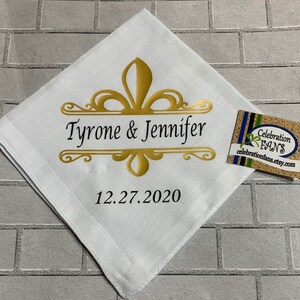 May include: White linen handkerchief with a gold fleur-de-lis design and the text "Tyrone & Jennifer" and "12.27.2020".