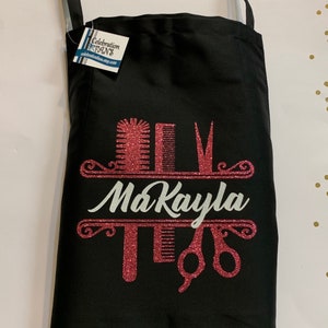 K&ouml;nnte beinhalten: Schwarze Sch&uuml;rze mit einem rosa Glitzerdesign mit K&auml;mmen, Scheren und dem Namen "MaKayla" in wei&szlig;er Schrift. Die Sch&uuml;rze hat einen Nackenriemen und ein Etikett von CelebrationFans.etsy.com.