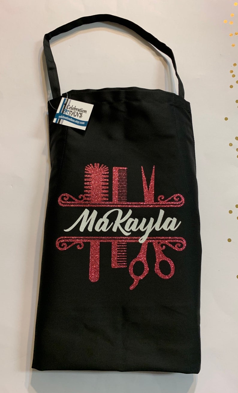 K&ouml;nnte beinhalten: Schwarze Sch&uuml;rze mit einem rosa Glitzerdesign mit K&auml;mmen, Scheren und dem Namen "MaKayla" in wei&szlig;er Schrift. Die Sch&uuml;rze hat einen Nackenriemen und ein Etikett von CelebrationFans.etsy.com.