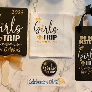 May include: A set of black and gold themed party favors for a girls trip to New Orleans. The set includes a hand fan, a door hanger, a button, and a cloth napkin. The hand fan has a gold ribbon and the text "2023 Girls Trip New Orleans". The door hanger has the text "Do Not Disturb Girls Trip New Orleans". The button has the text "Girls Trip". The cloth napkin has the text "Girls Trip".