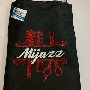 K&ouml;nnte beinhalten: Schwarze Sch&uuml;rze mit Schulterriemen. Die Sch&uuml;rze hat ein rotes Glitzerdesign mit dem Wort "Mijazz" in Wei&szlig; sowie Illustrationen von K&auml;mmen, Scheren und anderen Salonwerkzeugen. Ein Etikett ist an der Sch&uuml;rze befestigt.