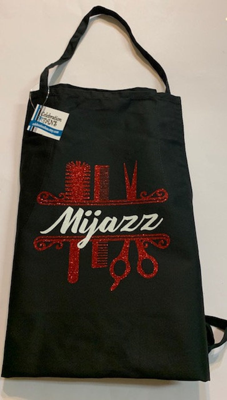 K&ouml;nnte beinhalten: Schwarze Sch&uuml;rze mit Schulterriemen. Die Sch&uuml;rze hat ein rotes Glitzerdesign mit dem Wort "Mijazz" in Wei&szlig; sowie Illustrationen von K&auml;mmen, Scheren und anderen Salonwerkzeugen. Ein Etikett ist an der Sch&uuml;rze befestigt.