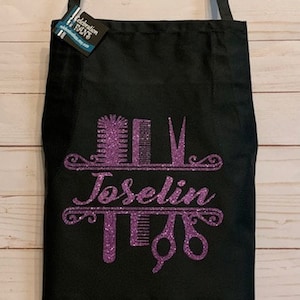 K&ouml;nnte beinhalten: Schwarze Sch&uuml;rze mit einem lila Glitzerdesign mit Friseurwerkzeugen und dem Namen "Joselin". Die Sch&uuml;rze hat einen Nackengurt und ein Etikett mit der Aufschrift "Celebration Days".