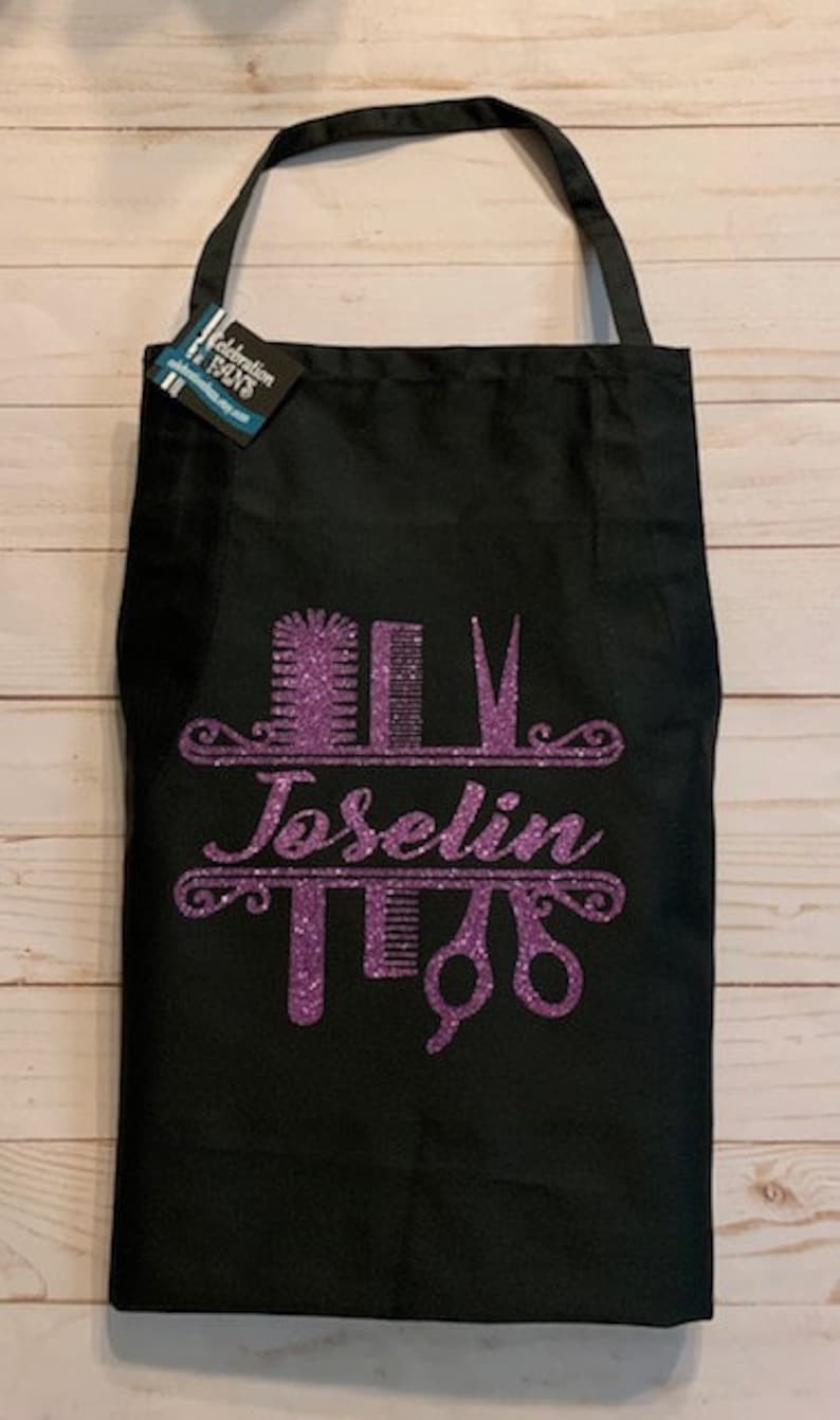 K&ouml;nnte beinhalten: Schwarze Sch&uuml;rze mit einem lila Glitzerdesign mit Friseurwerkzeugen und dem Namen "Joselin". Die Sch&uuml;rze hat einen Nackengurt und ein Etikett mit der Aufschrift "Celebration Days".