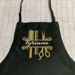 K&ouml;nnte beinhalten: Schwarze Sch&uuml;rze mit einem goldglitzernden Design mit Kamm, Schere und Haarb&uuml;rste. Der Name "Tyriane" ist in wei&szlig;er Schrift dargestellt. Die Sch&uuml;rze hat einen Nackenriemen und Bindeb&auml;nder.