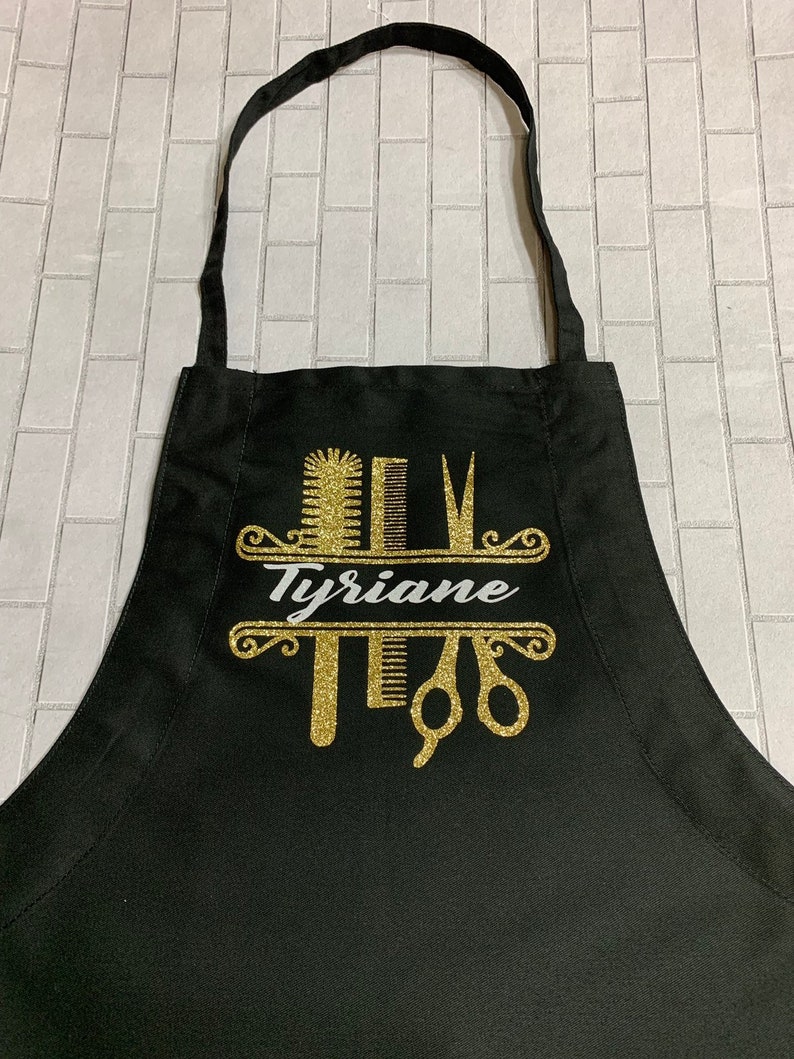 K&ouml;nnte beinhalten: Schwarze Sch&uuml;rze mit einem goldglitzernden Design mit Kamm, Schere und Haarb&uuml;rste. Der Name "Tyriane" ist in wei&szlig;er Schrift dargestellt. Die Sch&uuml;rze hat einen Nackenriemen und Bindeb&auml;nder.