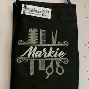 K&ouml;nnte beinhalten: Schwarze Sch&uuml;rze mit silbernem Glitzerdesign mit Friseurwerkzeugen und dem Namen "Markie". Die Sch&uuml;rze hat einen einzelnen Nackenriemen und ein Etikett von Celebration Fans.