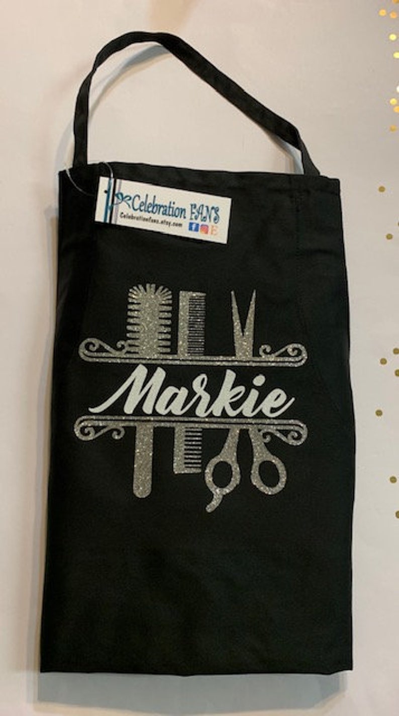 K&ouml;nnte beinhalten: Schwarze Sch&uuml;rze mit silbernem Glitzerdesign mit Friseurwerkzeugen und dem Namen "Markie". Die Sch&uuml;rze hat einen einzelnen Nackenriemen und ein Etikett von Celebration Fans.