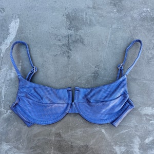 May include: A blue ribbed bikini top with adjustable straps and a clasp closure. The top has a structured, underwire design and a U-shaped neckline. The fabric has a subtle sheen, and the overall style is modern and minimalist.