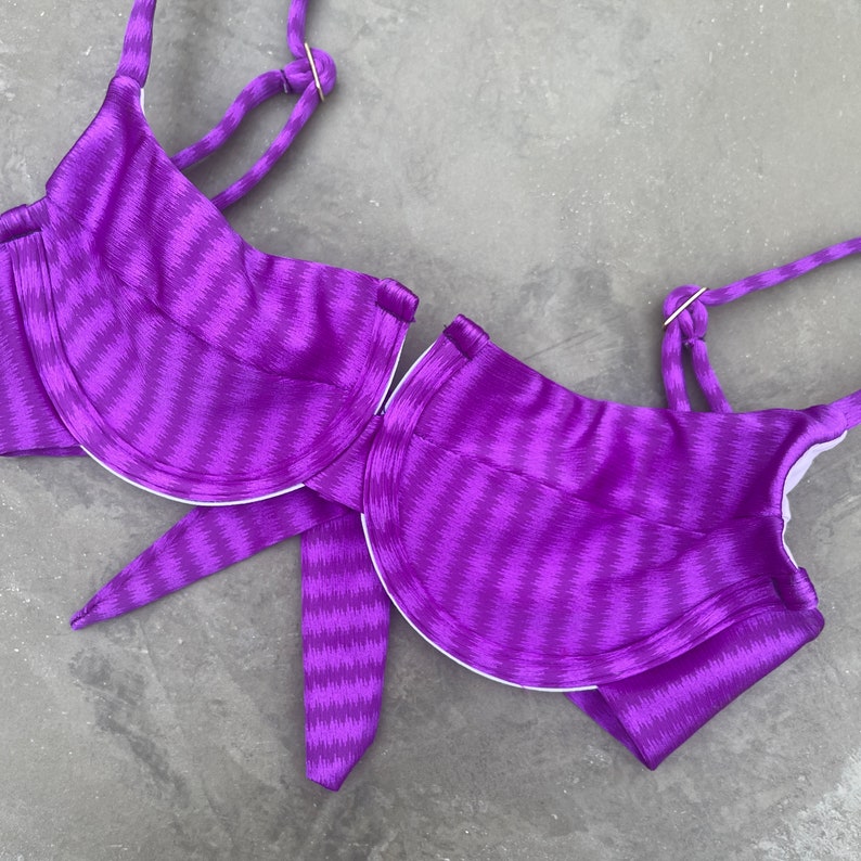May include: Purple bikini top with a striped pattern and underwire cups. The top has adjustable straps and a tie closure at the back.