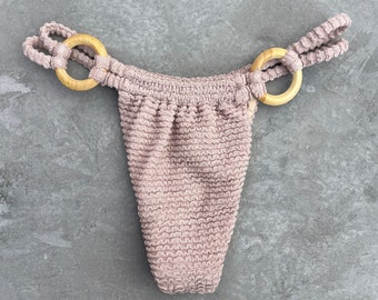 May include: A mauve-colored bikini bottom with a ruffled texture and two wooden rings on the sides.